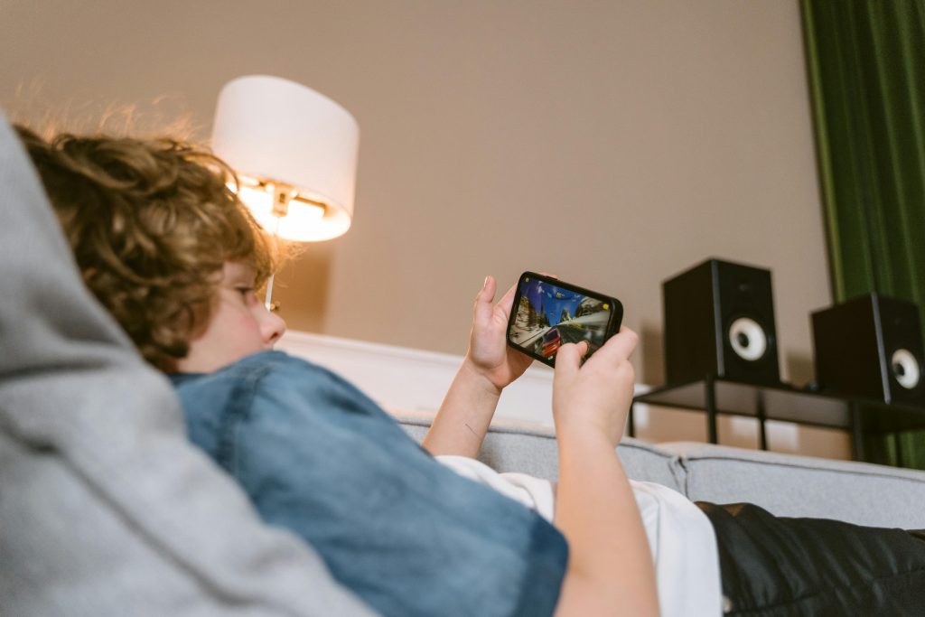 Teen lying on sofa playing video games on smartphone indoors, surrounded by modern décor.